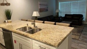 A kitchen with granite countertops, a double sink, and white cabinetry. There is a dining area with a table and chairs, and a living room with a couch and a TV. - One Block from Beach-Pelicans Landing! - Unit 1405 - Island Dunes Condo's (Port Aransas)