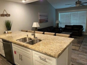 A kitchen with granite countertops, a double sink, and white cabinetry. There is a dining area with a table and chairs, and a living room with a couch and a TV.