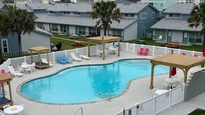 A swimming pool with a wooden deck, surrounded by white fences and various seating areas. - One Block from Beach-Pelicans Landing! - Unit 1405 - Island Dunes Condo's (Port Aransas)