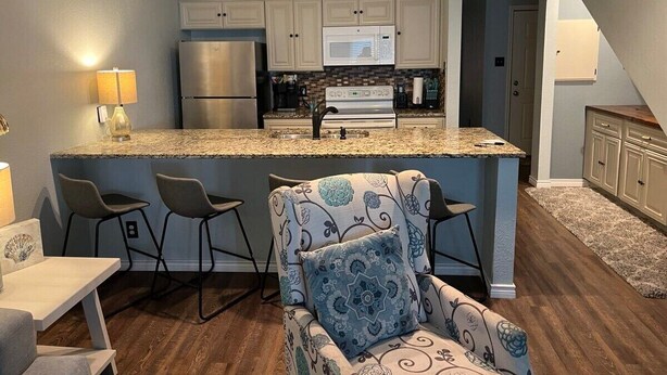 Cocina moderna con armarios blancos, un refrigerador de acero inoxidable, un microondas y encimera de granito. Hay tres taburetes de bar y un sillón estampado.