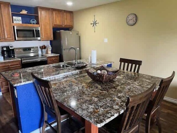 A kitchen with wooden cabinets, a microwave, a refrigerator, and a dining area with a granite countertop and wooden chairs.