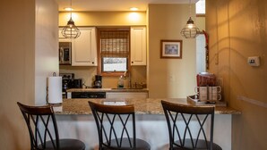 A kitchen with three bar stools, a microwave, a coffee maker, and a window with blinds.