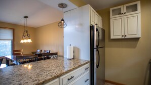A kitchen with granite countertops, white cabinets, a stainless steel refrigerator, and a dining area with wooden chairs and a table.