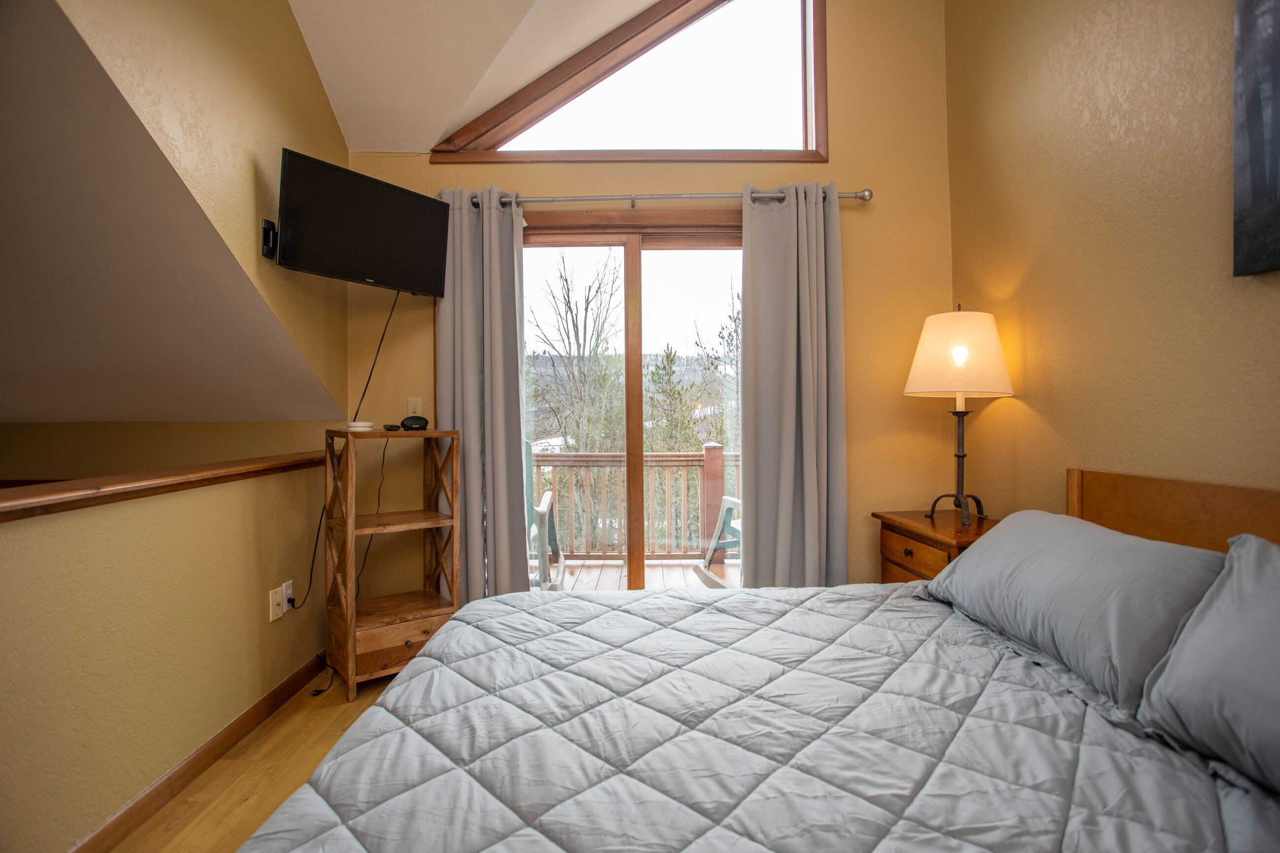 A bedroom with a bed, a TV mounted on the wall, a nightstand with a lamp, and a balcony with a view.