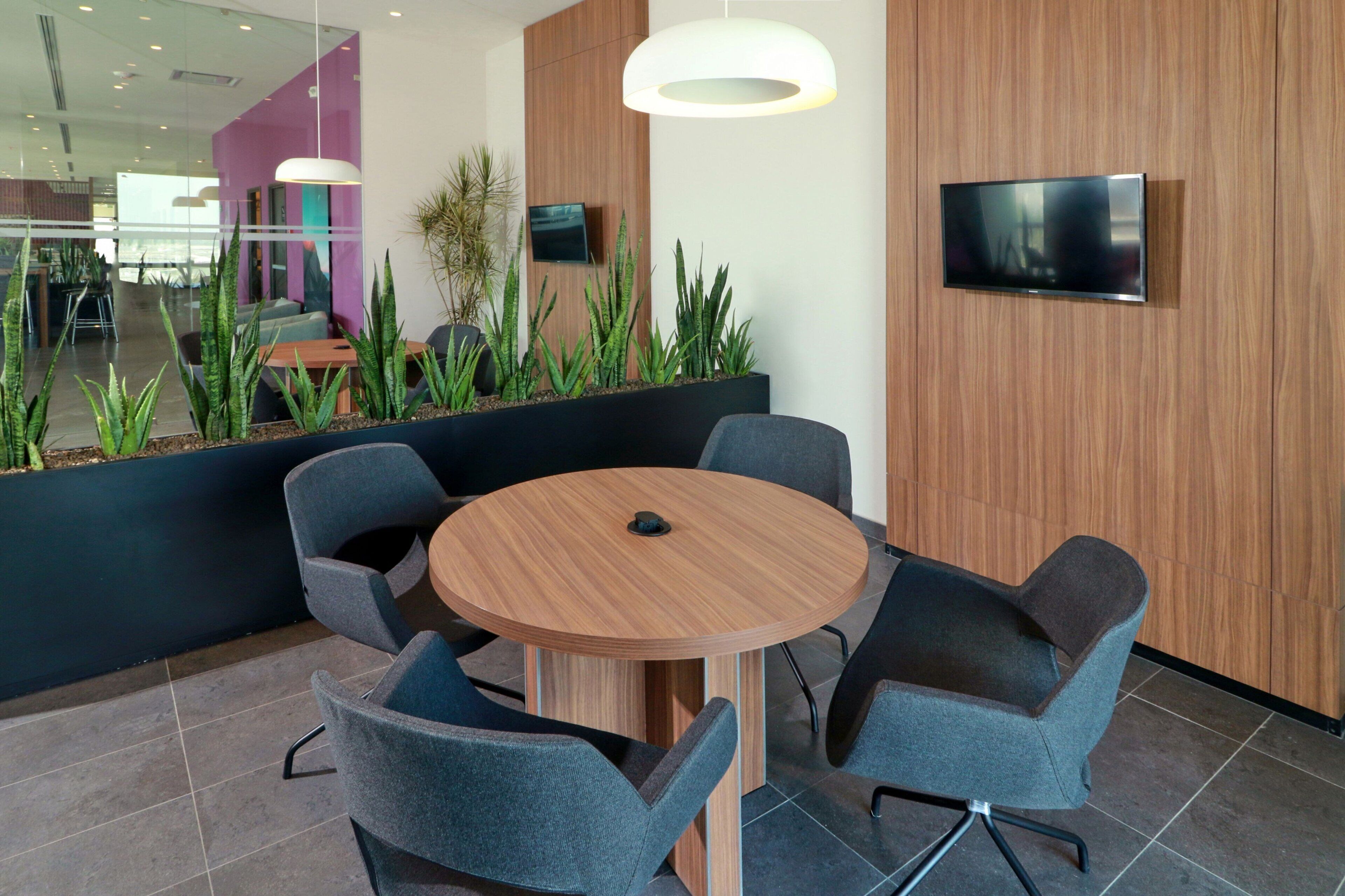 Un espace de bureau moderne avec une table ronde en bois, entourée de quatre chaises grises, d’un canapé noir et d’un téléviseur fixé au mur.