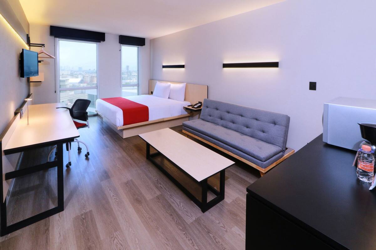 Habitación de hotel moderna con cama, sofá, escritorio y un amplio ventanal con vistas a la ciudad.