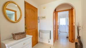 Un pasillo con una puerta de madera, un teléfono blanco montado en la pared, un radiador blanco, un armario blanco con una cesta y un jarrón con plantas secas.