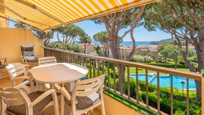 Un balcón con una mesa y sillas blancas, un dosel a rayas y vistas a una piscina y árboles.