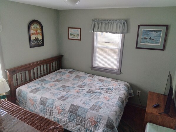 A bedroom with a wooden bed frame, a quilted bedspread, a window with blinds, and a wall-mounted clock.