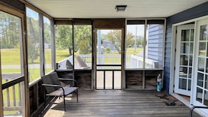 A covered porch with a bench, a table, and a view of a house and trees. - Shucker's Tump -  pet friendly, Lg screened porch. LINENS AND TOWELS PROVIDED (Chincoteague)