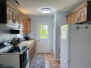 A kitchen with wooden cabinets, a white refrigerator, a stove, and a window with a view of trees. - Shucker's Tump -  pet friendly, Lg screened porch. LINENS AND TOWELS PROVIDED (Chincoteague)
