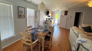 A dining area with a wooden table, chairs, and a chandelier. There is a kitchen in the background with cabinets and a refrigerator. - Shucker's Tump -  pet friendly, Lg screened porch. LINENS AND TOWELS PROVIDED (Chincoteague)