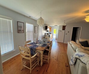 A dining area with a wooden table, chairs, and a chandelier. There is a kitchen in the background with cabinets and a refrigerator. - Shucker's Tump -  pet friendly, Lg screened porch. LINENS AND TOWELS PROVIDED (Chincoteague)