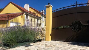Une entrée pavée de pierres, avec un portail en métal, un bâtiment jaune doté d’un toit rouge et un jardin agrémenté de lavande.