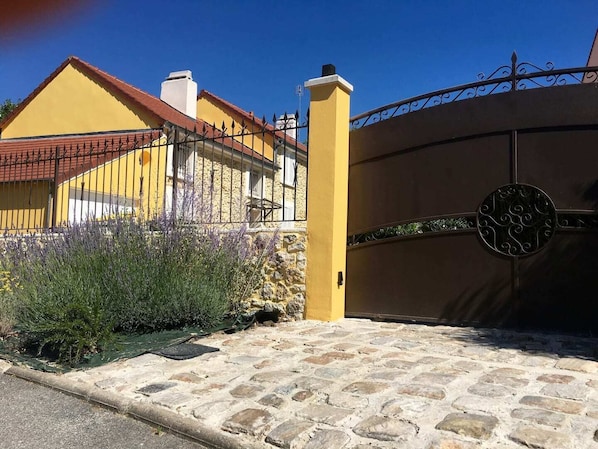 A stone-paved entrance with a metal gate, a yellow building with a red roof, and a garden with lavender. - Beautifully renovated studio between Paris and Disneyland (Marolles-en-Brie)