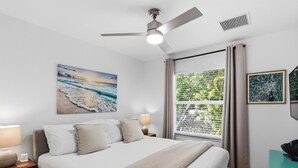 Ein Schlafzimmer mit einem Bett, einem Deckenventilator, einem Fenster mit Vorhängen und Kunstwerken an den Wänden.
