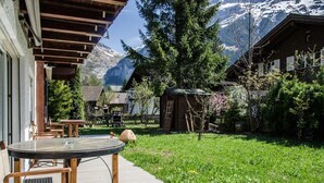 A wooden cabin with a glass table and chairs, surrounded by a lush garden and a mountain backdrop.