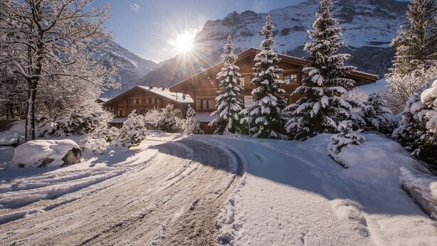 A snow-covered mountain landscape with a wooden lodge, a clear sky, and a sun shining through the trees.