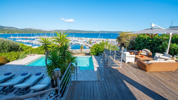 Een houten terras met lounge stoelen en een zwembad, met uitzicht op een jachthaven met boten en een helderblauwe lucht.