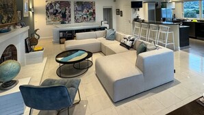 Een moderne woonkamer met een grote hoekbank, een ronde salontafel en een blauwe fauteuil. Er is ook een keuken met een bar en zitplaatsen.
