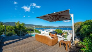 Een houten terras met rieten meubilair, een tafel en een parasol, met uitzicht op een jachthaven en heuvels.