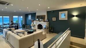 Een moderne woonkamer met een open haard, een eetgedeelte met uitzicht op zee en een trap naar een hoger niveau.