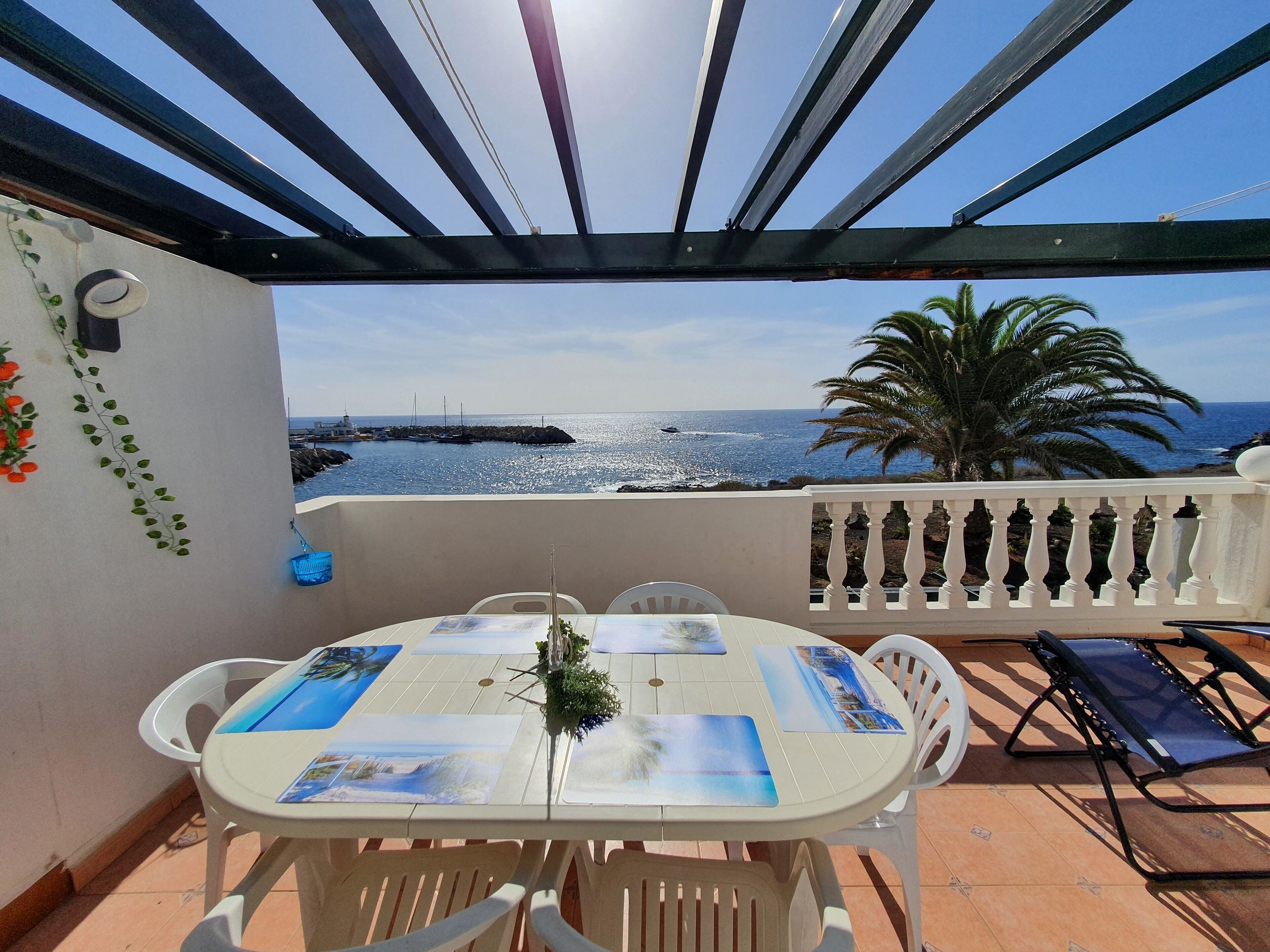 Une terrasse avec une table dressée pour deux personnes, offrant une vue sur la côte, avec un brise-lames rocheux et des palmiers.