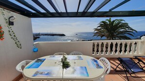 A terrace with a table set for two, overlooking a coastal view with a rocky breakwater and palm trees.