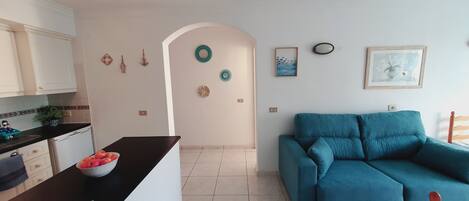 A modern living room with a blue sofa, a kitchen area with white cabinets, and decorative wall art.