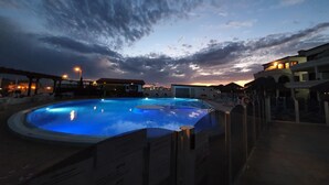 A swimming pool with a sunset view, surrounded by buildings and a covered area with lounge chairs.