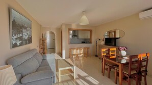 Una sala de estar con un sofá, una mesa de centro y un televisor. Se observa un comedor con una mesa y sillas. Se puede ver una cocina al fondo.