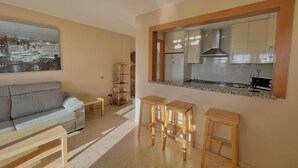Un espacio habitable funcional con sofá, pequeña mesa de comedor y tres taburetes. Se ve una cocina con armarios y encimera.
