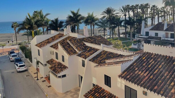 Un edificio costero con techos de tejas terracota, paredes blancas y vista a la playa y palmeras.