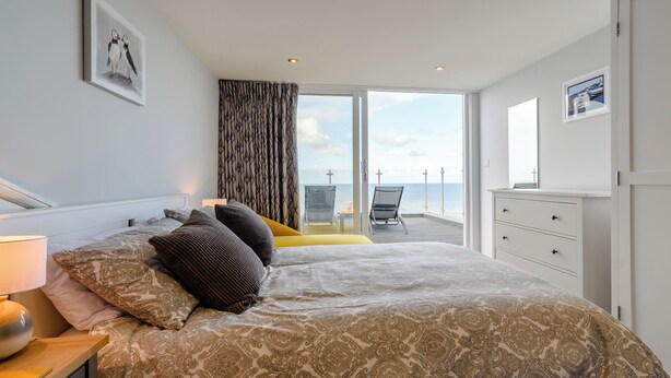 A bedroom with a large bed, a nightstand, a dresser, and a view of the sea through a sliding glass door. - 3 bedroom accommodation in Reighton Gap, near Filey (Reighton Gap, near Filey)