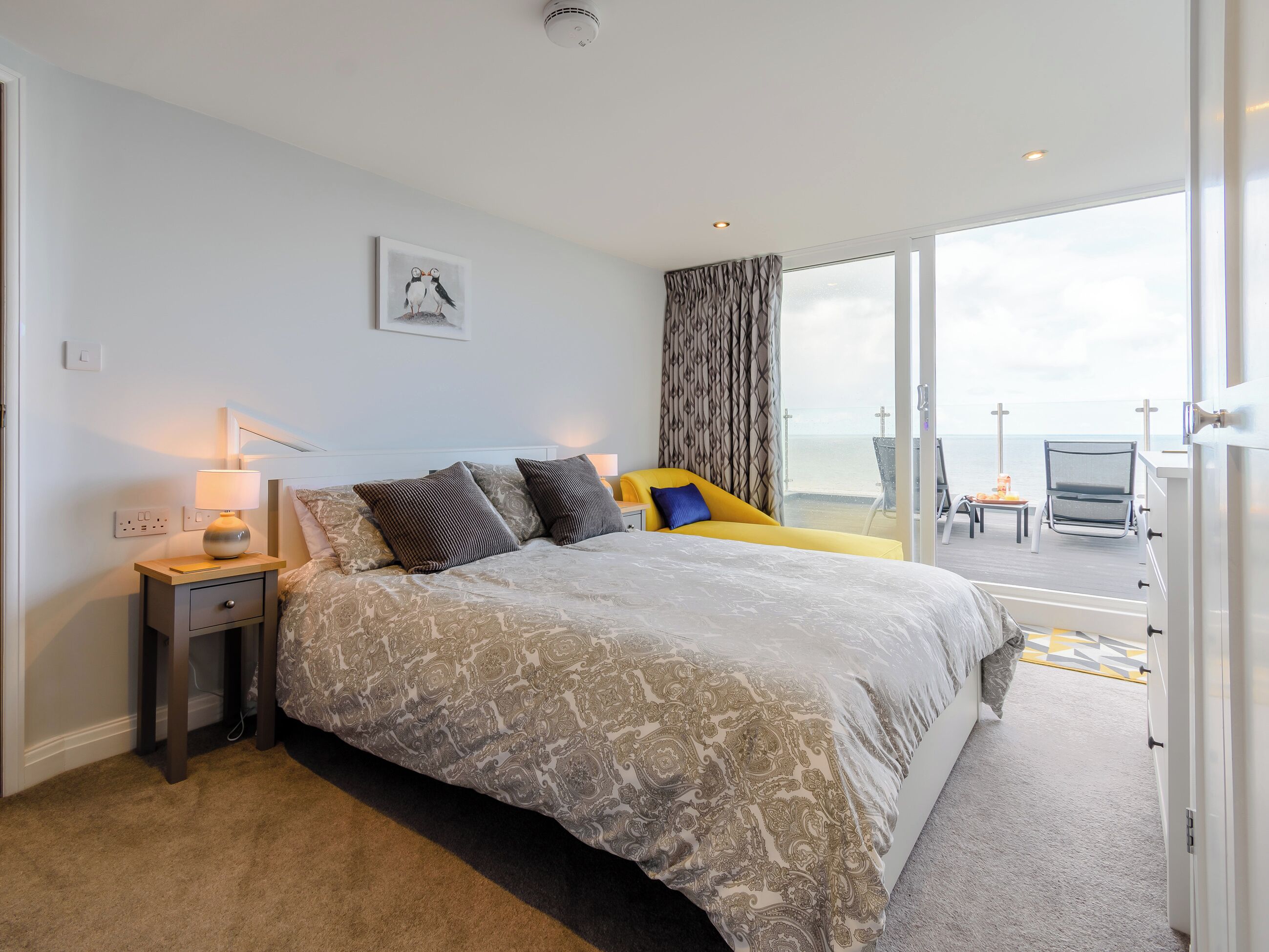 A hotel room with a large bed, a bedside table with a lamp, a view of the sea through a large window, and a balcony with a chair.