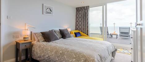 A hotel room with a large bed, a bedside table with a lamp, a view of the sea through a large window, and a balcony with a chair.