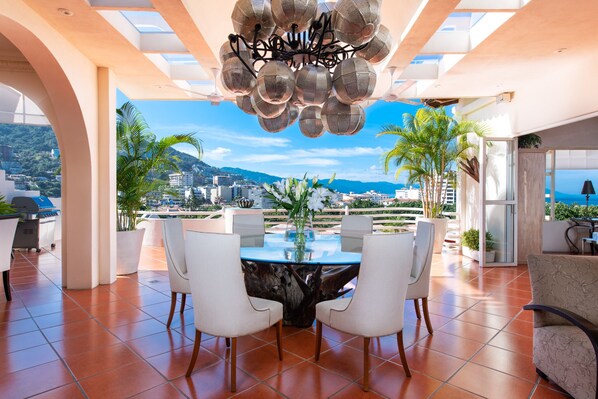 A modern dining area with a glass table, white chairs, and a view of the city and mountains.