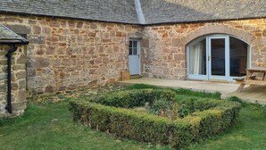 Un edificio de piedra con techo de tejas, una ventana pequeña y una puerta.  Hay una terraza de madera con un banco y un seto bien cuidado al frente.