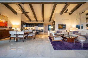 Ampla sala de estar com um conjunto de jantar, sofá e mesa de centro. Há ventiladores de teto, uma TV e uma cozinha ao fundo.