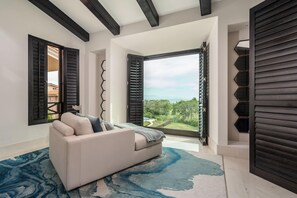 Uma sala de estar moderna com sofá, vista para vegetação e uma grande janela com persianas.
