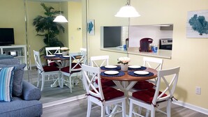 A dining area with a round table, white chairs, and red cushions. There is a television on a stand, a plant, and a mirror reflecting the room. - SABTR- 710- 6 MIN. WALK TO THE BEACH! POOL, CLEAN,2BR,2BA VILLA, 5-STAR REVIEWS! (St. Augustine)