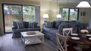 A living room with a grey sofa set, a white coffee table, and a dining area with a wooden table set for two. - SABTR- 710- 6 MIN. WALK TO THE BEACH! POOL, CLEAN,2BR,2BA VILLA, 5-STAR REVIEWS! (St. Augustine)