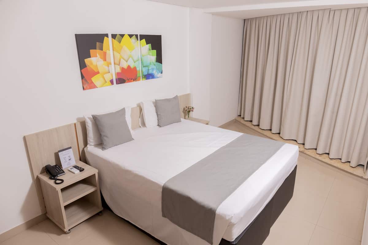 Ein Hotelzimmer mit einem großen Bett, einem Nachttisch mit Telefon und Lampe, einem an der Wand hängenden Kunstwerk und einem Fenster mit Vorhängen.
