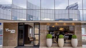 Une entrée d’hôtel moderne avec une façade en verre, un intérieur visible avec des sièges, et de grandes plantes en pot disposées à l’entrée.