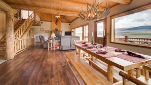 Un intérieur rustique avec un escalier en bois, une table à manger recouverte de nappes à carreaux et une grande fenêtre donnant sur l’extérieur.