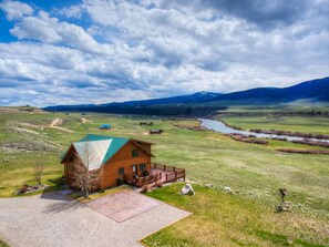 A wooden cabin with a green roof, surrounded by a grassy landscape, a river, and mountains in the background.