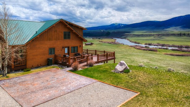 Une cabane en bois avec un toit vert, une terrasse et une vue sur une rivière et des montagnes.