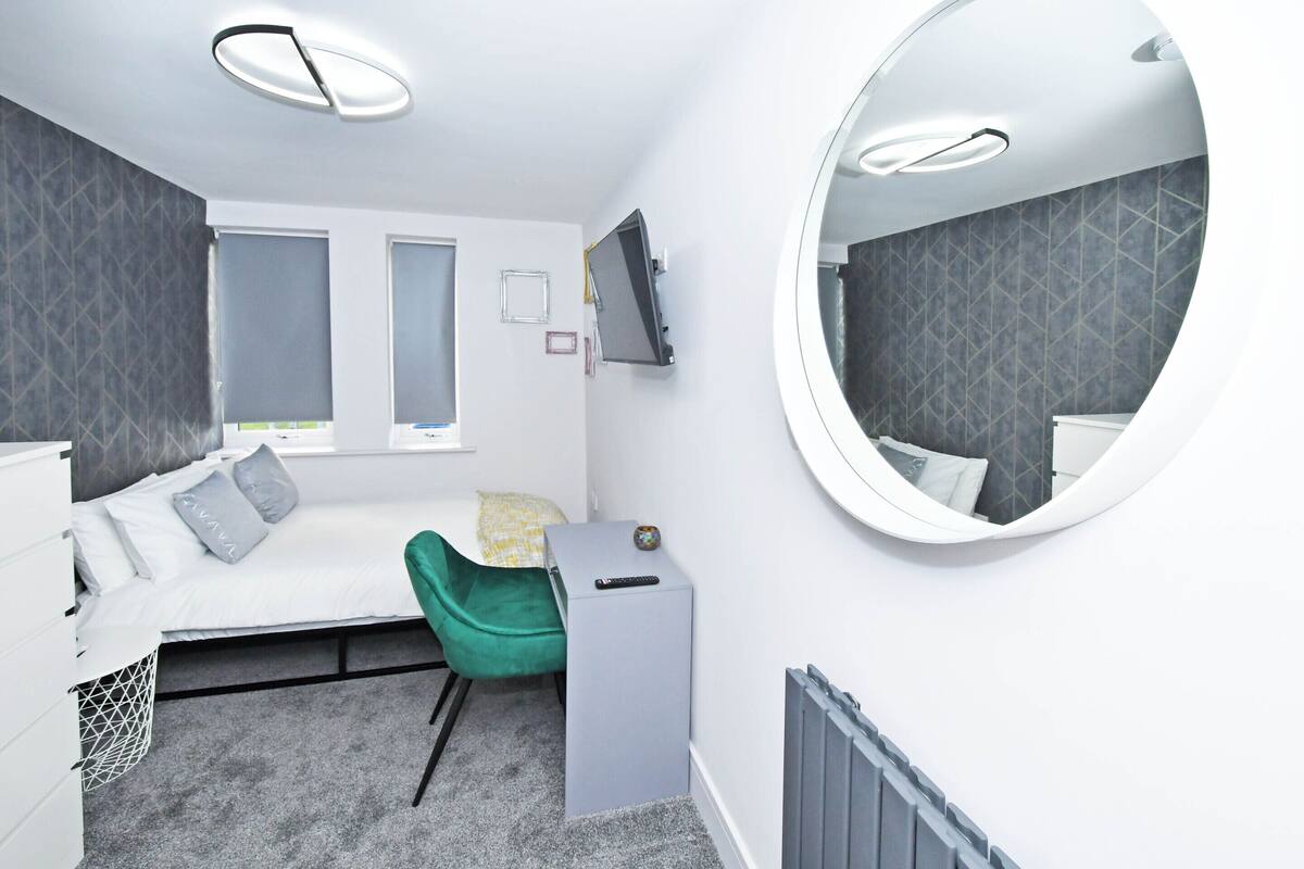 Un dormitorio moderno con cama, escritorio, silla, televisor y un espejo grande.
