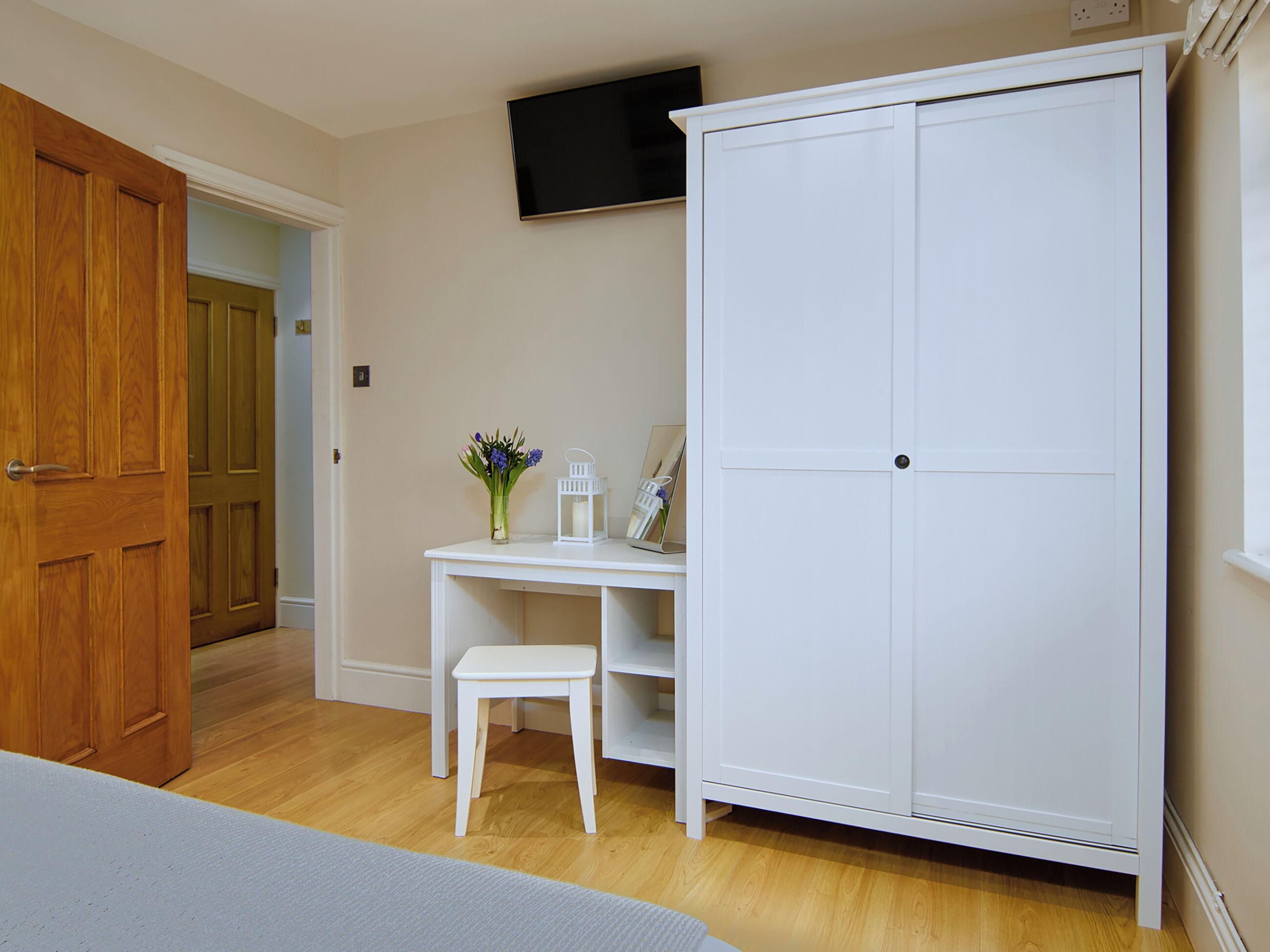 A bedroom with a wooden door, a white wardrobe, a desk with a chair, and a television on the wall.