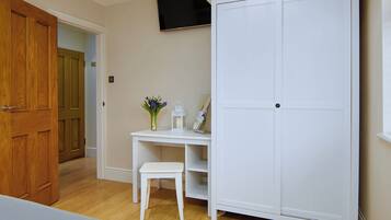 A bedroom with a wooden door, a white wardrobe, a desk with a chair, and a television on the wall.